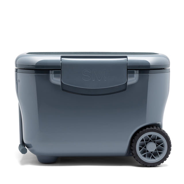 Gray-blue hard-sided rolling cooler with hinged lid, wheels, pull handle, and embossed SM logo, photographed against neutral background. Gray-blue hard-sided rolling cooler with hinged lid, wheels, pull handle, and embossed SM logo, photographed against neutral background.
