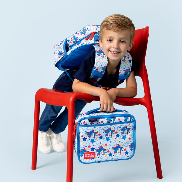 Young boy smiling at camera, sitting on red plastic chair wearing navy polo shirt, white star-patterned bandana, and white sneakers. Holding matching star-print lunch box with blue trim and SM logo. Drawstring bag visible