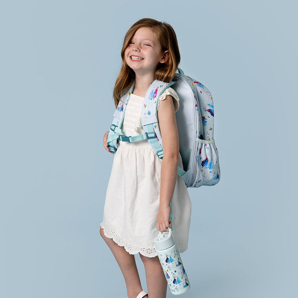 Young girl wearing white dress with blue and purple printed jacket, holding matching backpack and water bottle with geometric and nature designs, white sneakers, against blue-gray background.