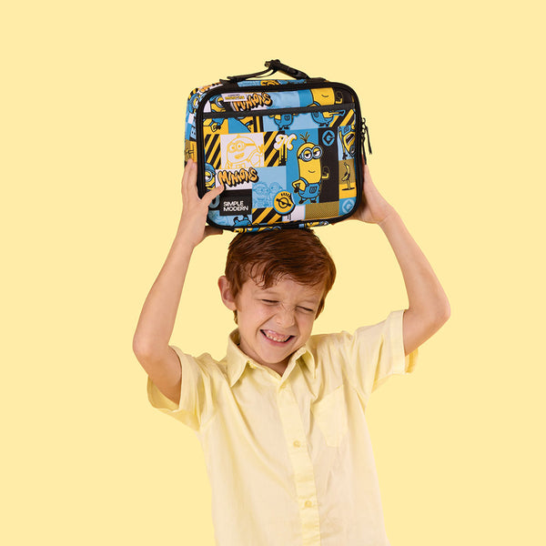 Young boy with reddish-brown hair wearing a yellow shirt holds a Simple Modern Minions lunch bag above his head, smiling with joy against a pale yellow background.