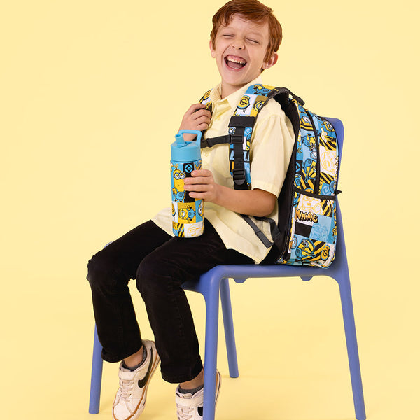 Young boy with reddish-brown hair smiling on a blue chair, wearing cream shirt and black pants, holding a blue Simple Modern water bottle and wearing a matching Minion-patterned backpack against a pale yellow background.
