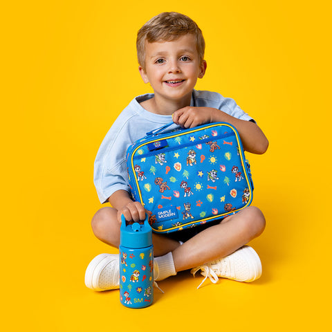Young boy smiling while holding a blue Paw Patrol lunch bag and water bottle with matching cartoon character designs, against a bright yellow background.