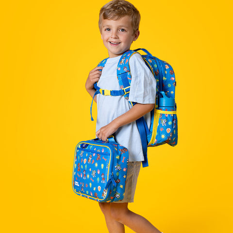Young boy smiling at camera wearing light blue shirt, holding blue Paw Patrol backpack and lunch box with yellow trim against bright yellow background.