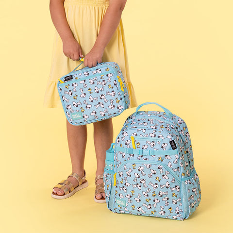 Two light blue Simple Modern bags featuring Peanuts characters on a pale yellow background. One bag is held by a person in a yellow dress; the second sits on the ground. Both have turquoise straps and yellow zippers.