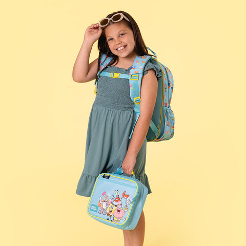 Young girl smiling at camera wearing sage green dress, holding light blue SpongeBob SquarePants lunch box and wearing matching backpack with yellow buckles against pale yellow background.