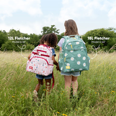 Two children standing back-to-back in a grassy field wearing Simple Modern Fletcher backpacks in pink and sage green, showing size comparison between 12L and 18L models.